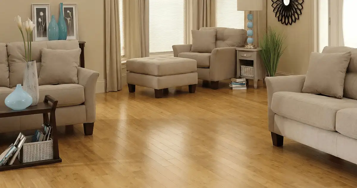 A living room with professionally installed bamboo flooring, beige sofas, a matching ottoman, a side table with a blue lamp, vases, framed pictures, and large windows with blinds and curtains in Cape Coral, FL